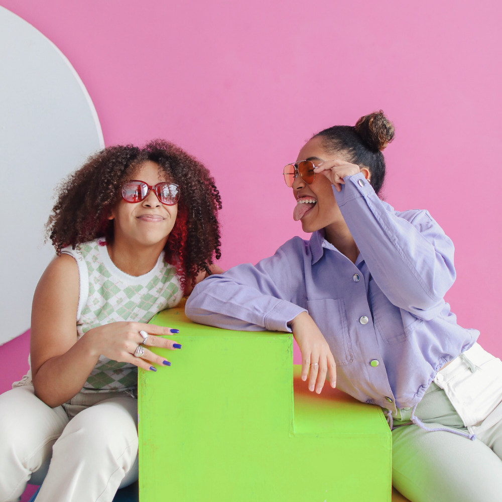 Two women sitting on a green cube against a pink background